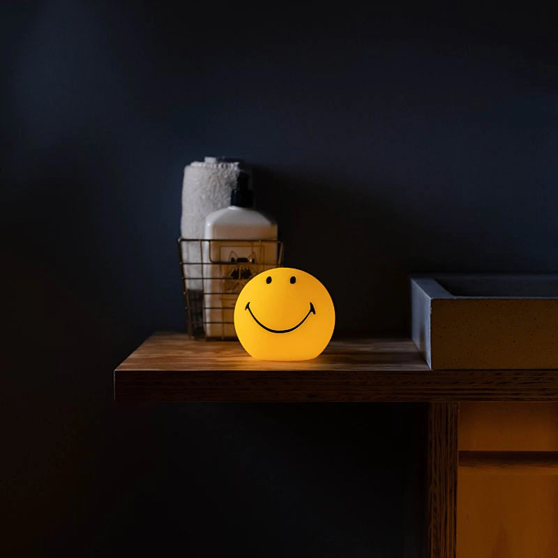 A cordless, round lamp with a yellow silicone body and a smiling face design on a wooden table in front of a dark navy wall.