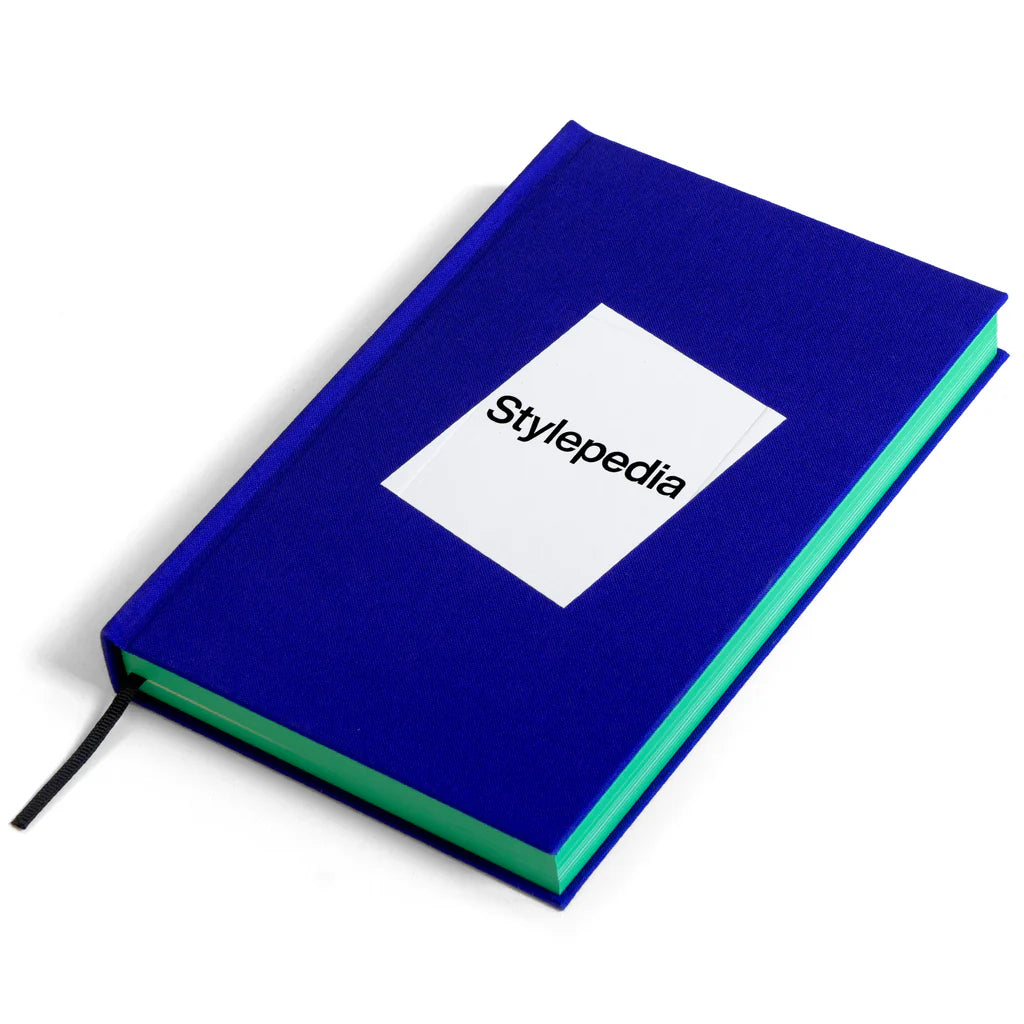 The front cover of 'Stylepedia' featuring the title inside a white tilted square against a dark blue background. The book is lying flat on a white surface at an angle and has green pages.