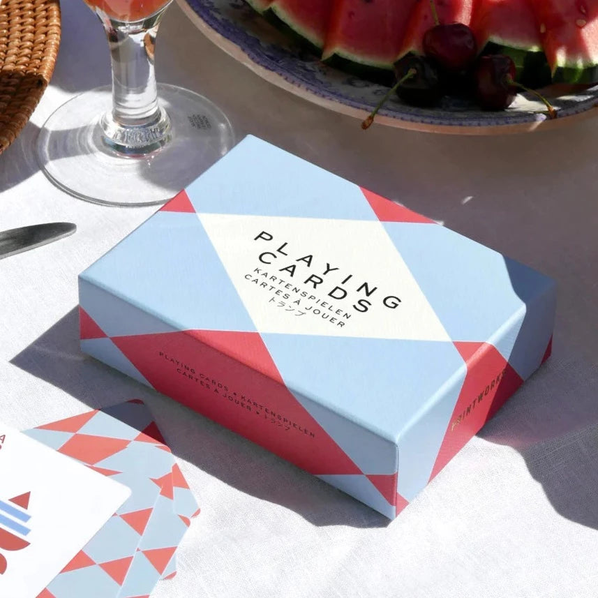 A red and blue box of playing cards on a table in the sun next to a glass and a bowl of watermelon.