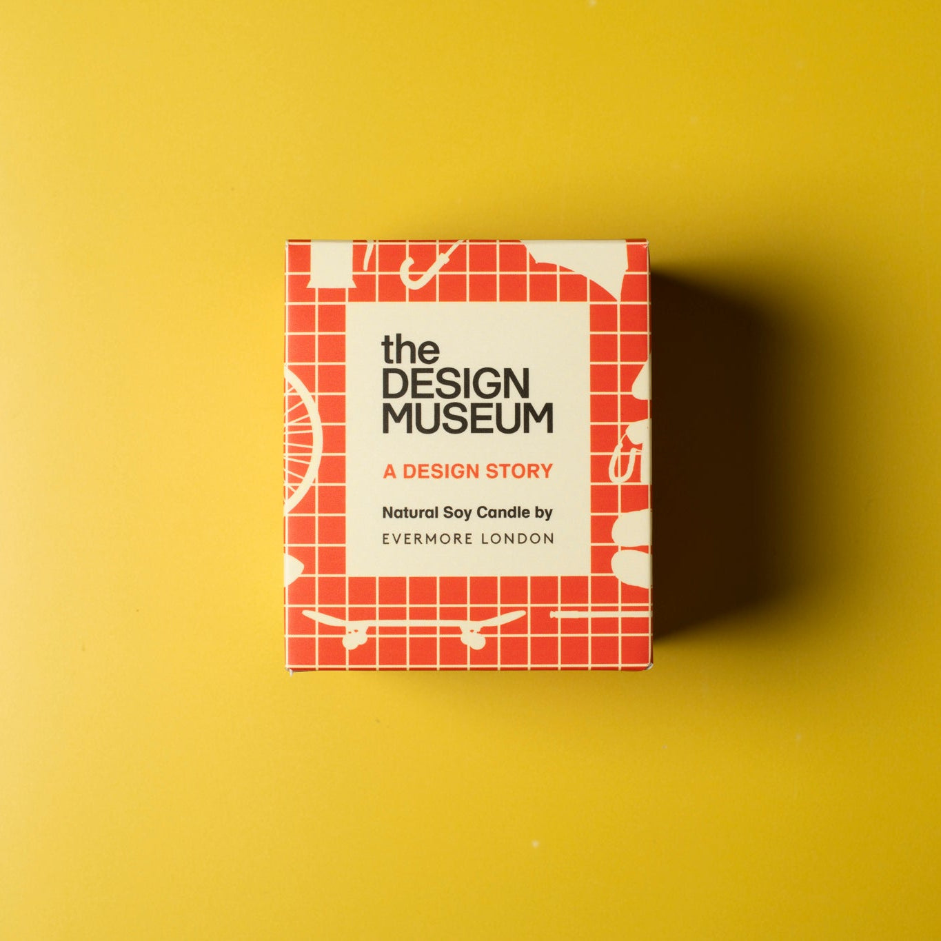 Orange grid design packaging box for the Design Museum candle on a yellow background. 