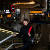Person standing on an escalator in London Bridge station, holding a black tote bag with a clock and digital displays in the background.