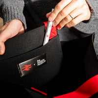 Person holding open a black tote bag with a visible brand logo, wearing a gray sweater.