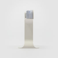 A minimalist white metal magazine rack with a curved pedestal base and a top shelf holding a stack of magazines, set against a seamless white studio background.