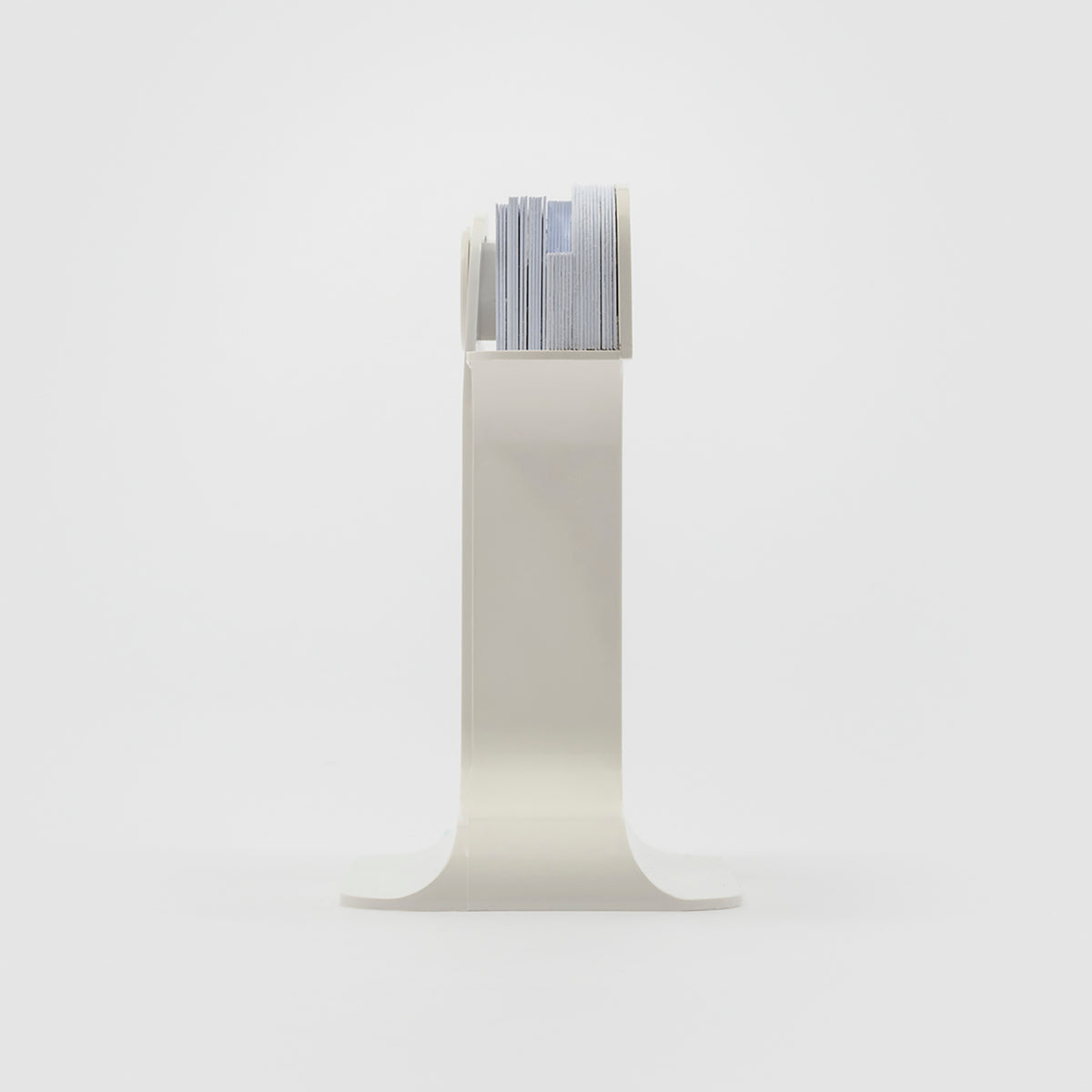 A minimalist white metal magazine rack with a curved pedestal base and a top shelf holding a stack of magazines, set against a seamless white studio background.