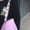 Person wearing a black shirt with graphic design and pink suspenders with pins, standing against a blurred background.