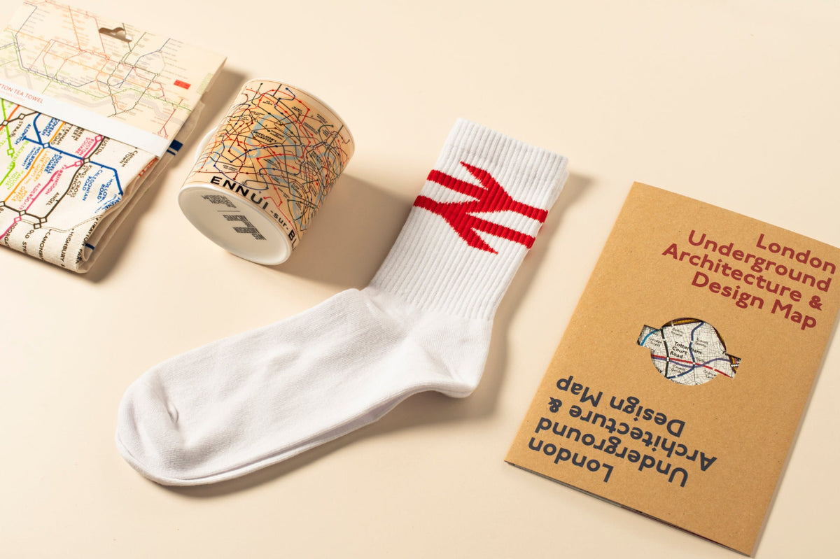 A white rail logo crew sock displayed on a beige surface alongside a London Underground Architecture & Design Map booklet, a map tea towel, and map mug