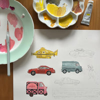 Art supplies including paint, brushes, and a sheet of colored car illustrations on a wooden surface.