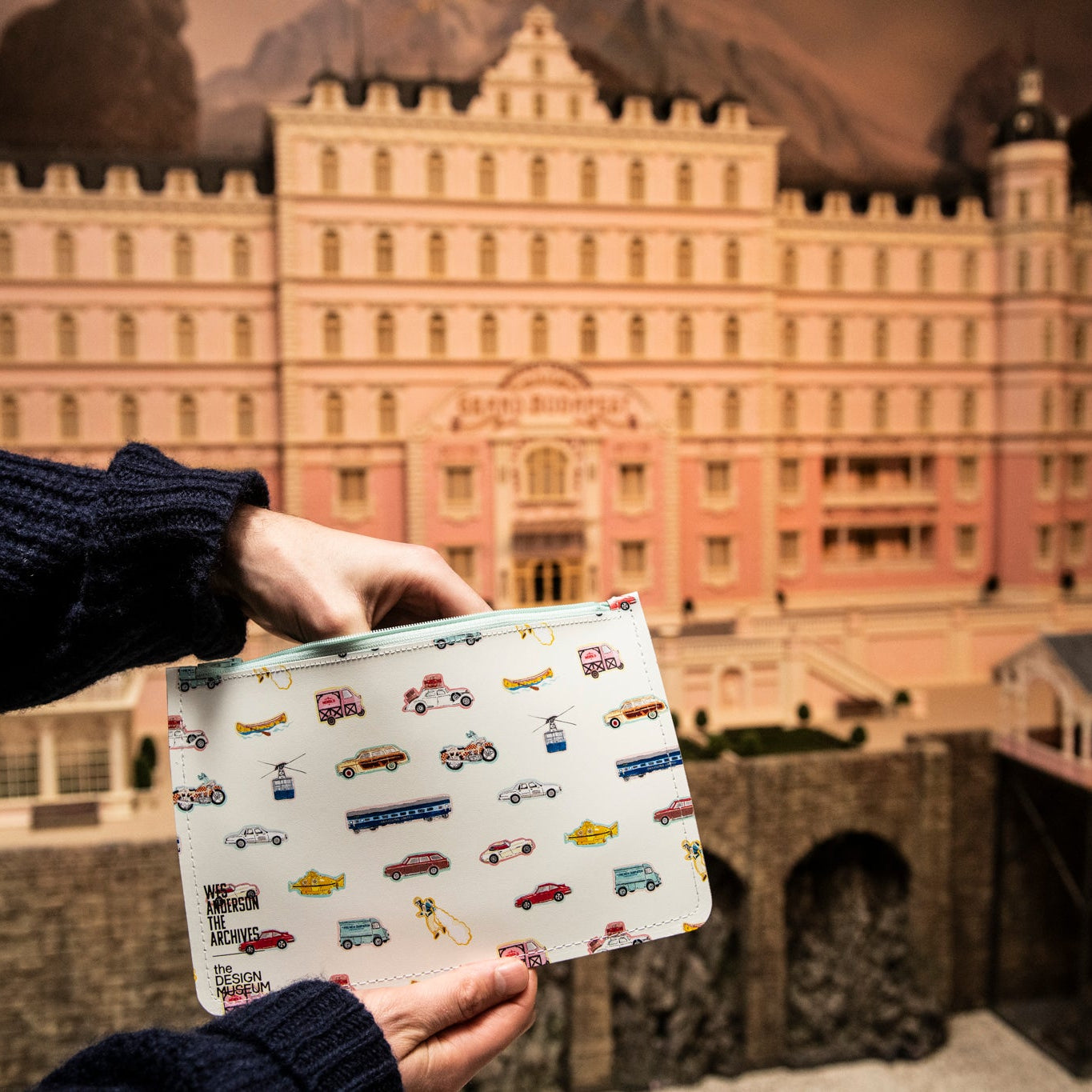 Person holding a white leather pouch with vehicle illustrtaions in front of a large building model.