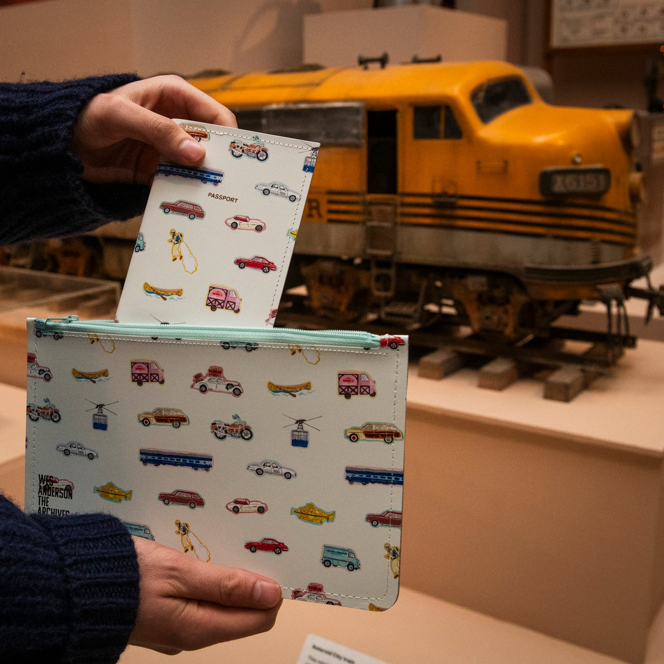 Person holding a white passport holder and pouch with vehicle illustrations in front of a model train set.