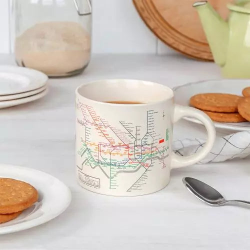 A ceramic mug with a white background featuring a vintage print of London's underground map in colorful lines on a white table with plates of biscuits.