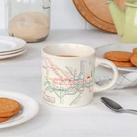 A ceramic mug with a white background featuring a vintage print of London's underground map in colorful lines on a white table with plates of biscuits.