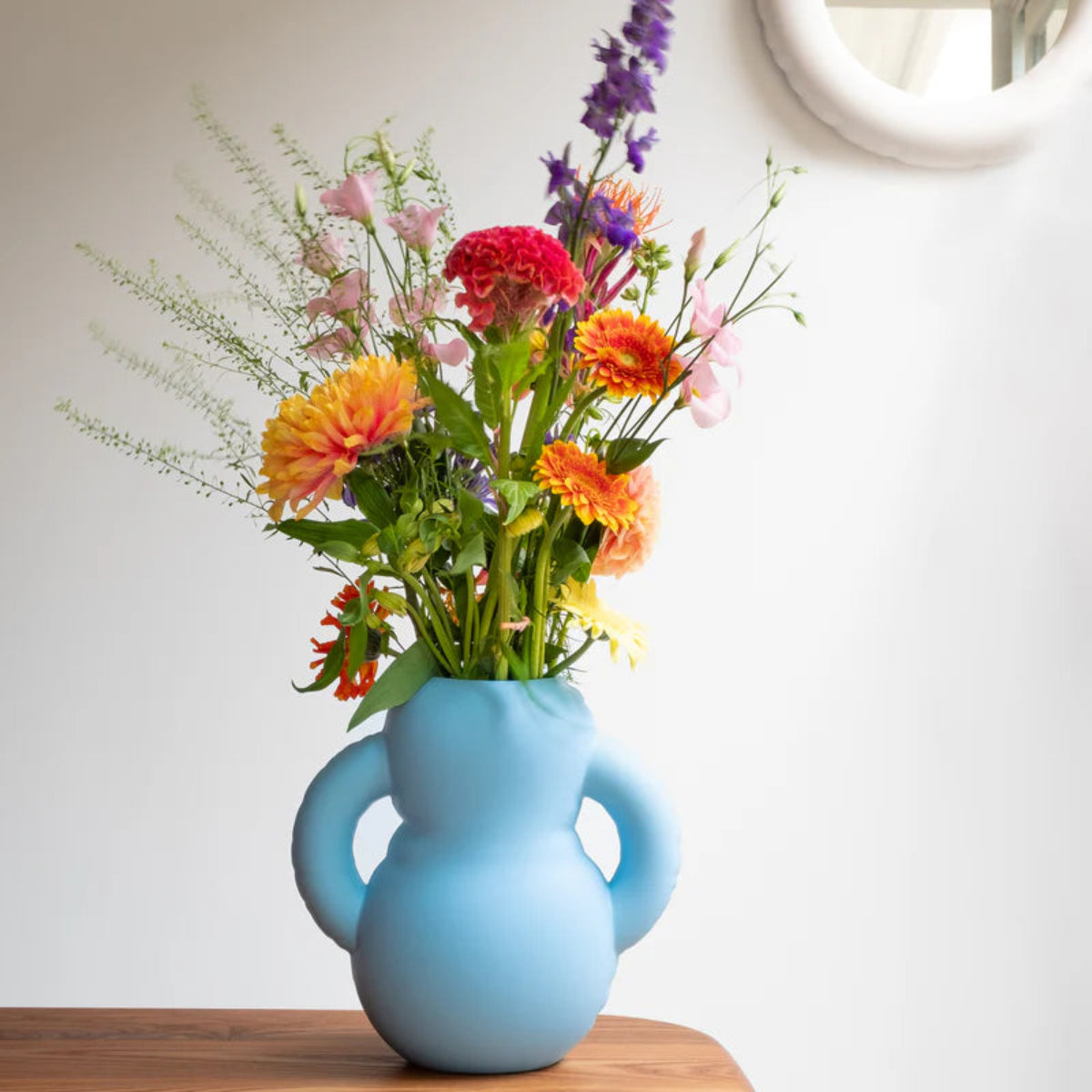 A light blue vase with colorful flowers on a wooden surface against a white wall.