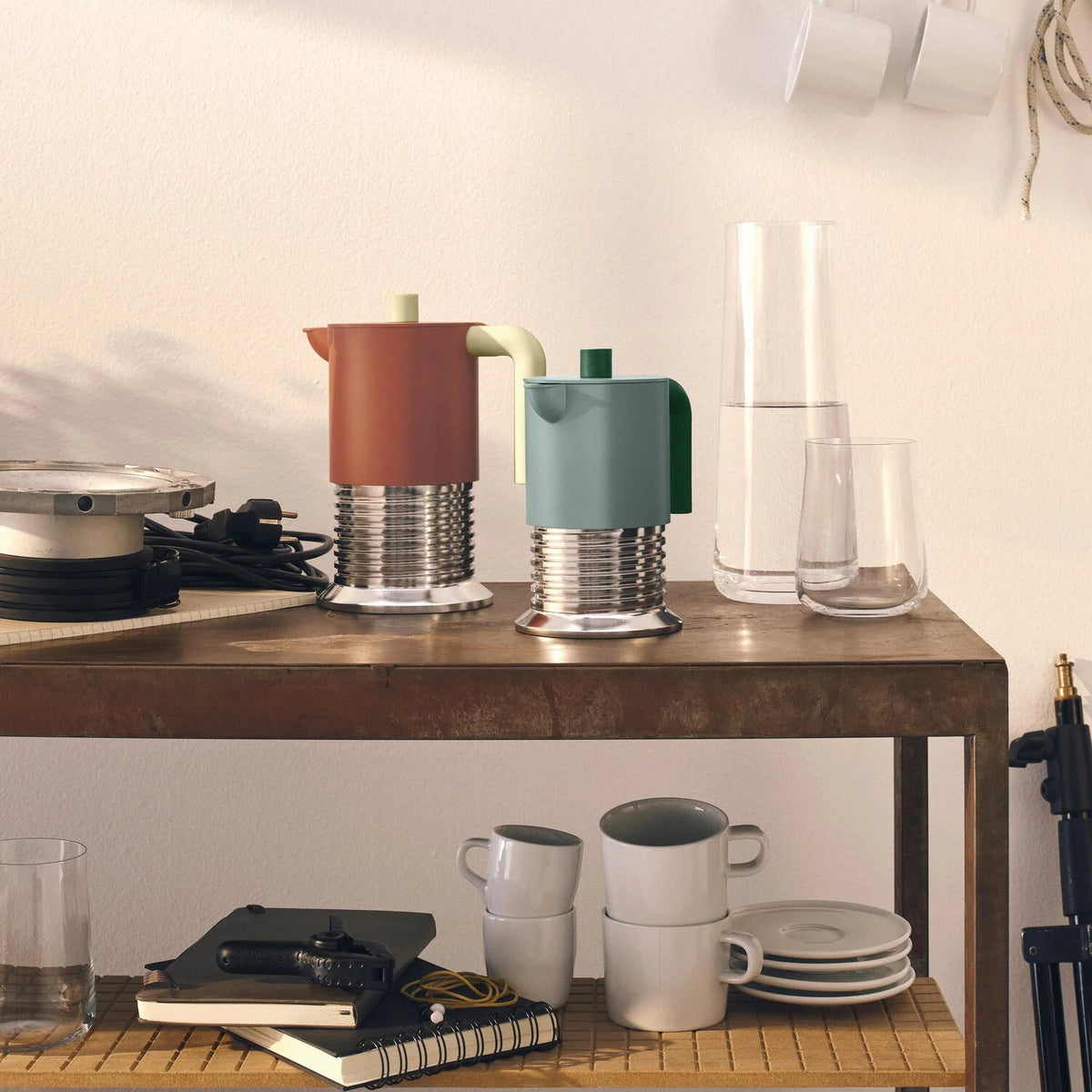 Two retro-style coffee makers with metallic bases, one terracotta orange and one sage green, displayed on a wooden shelf in a warm, minimalist interior setting.