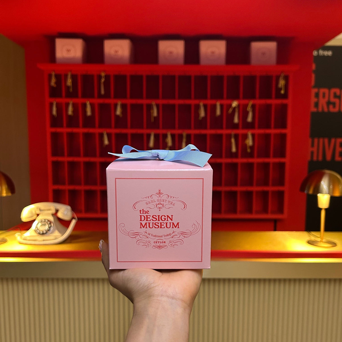 Pink Mendl's teabags box a blue ribbon with held in front of a red and gold reception desk