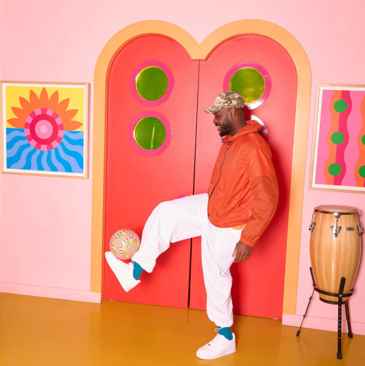 Person in orange jacket and white pants kicking a colourful football in front of a pink wall.