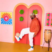 Person in orange jacket and white pants kicking a colourful football in front of a pink wall.